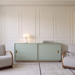 A light sage green two-door cabinet with gold handles in a classic room setting, flanked by two cream upholstered armchairs with curved wooden arms, set against a white paneled wall.