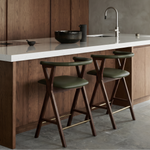 A modern kitchen island with a white countertop, dark wood cabinetry, a matte gray concrete wall, and two wooden bar stools with olive green seats in the foreground.