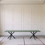 A long rectangular bench with a muted sage green top and black X-shaped metal legs, set against an elegant white paneled wall on a patterned rug.