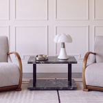A symmetrical interior scene with two upholstered armchairs facing a small dark marble side table with a white table lamp. The room has cream paneled walls, a light rug, and a dark wood shelving unit on the right.