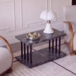 A modern two-tier side table with a dark gray marble top and base, supported by black metal legs, styled in a living room setting with a white mushroom-shaped table lamp and a small decorative tray on top.