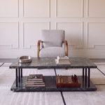 A modern two-tier coffee table with a dark green marble top and lower shelf, styled with books and a chess set, positioned in front of a light gray armchair against a cream paneled wall.