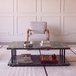 A modern two-tier coffee table with a dark gray marble top and base, black metal legs, and a wooden lower shelf, styled in a living room setting with books, a tray, and a chess set. A light gray armchair is positioned behind the table against a cream paneled wall.