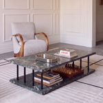 A modern two-tier coffee table with a dark green marble top and bottom shelf, styled with books, a chess set, and a metal tray. It is set in a bright, elegant room next to a light gray armchair with curved wooden arms.