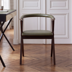 A modern armchair with a dark wood frame and an olive green upholstered seat and backrest, positioned in a classic room with white paneled walls and a wooden herringbone floor next to a black desk.