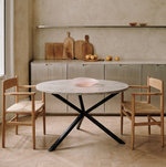 A dining table with a round white marble top and black metal X-base, paired with two light wood chairs with cream cushions, set in a minimalist dining room with a textured beige wall, wooden cutting boards, and a pendant light.