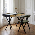 A modern writing desk with a black top, brushed gold metal apron, and black X-style legs, paired with a matching dark green upholstered chair in a bright, classic Parisian-style room with white paneled walls and herringbone wood floors.